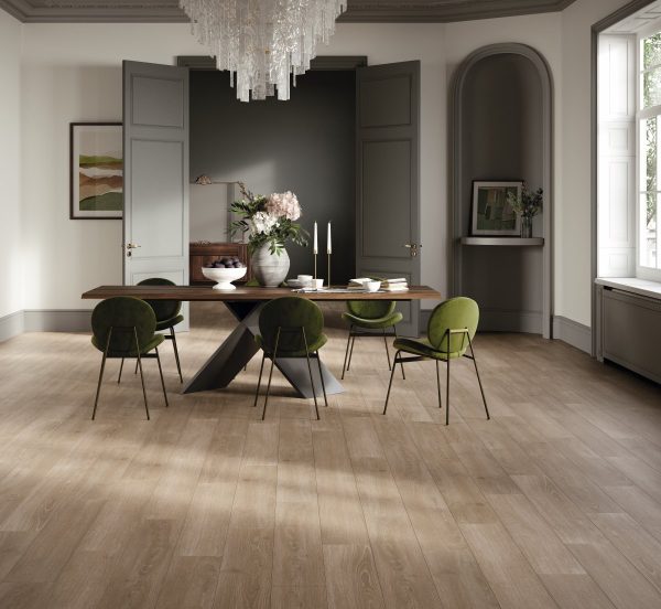 Ruime eetkamer met een eettafel en stoelen. Aan het plafond hangt een kristallen kroonluchter. Op de achtergrond zie je openslaande deuren naar de volgende ruimte. er ligt een mooie natuurlijk bruin/grijze laminaatvloer met waterbestendige toplaag van Parador in de ruimte. Type Parador Hydron 600, eiken skyline parelgrijs. In de gebruiksklassen van laminaat valt deze vloer onder klasse 32
