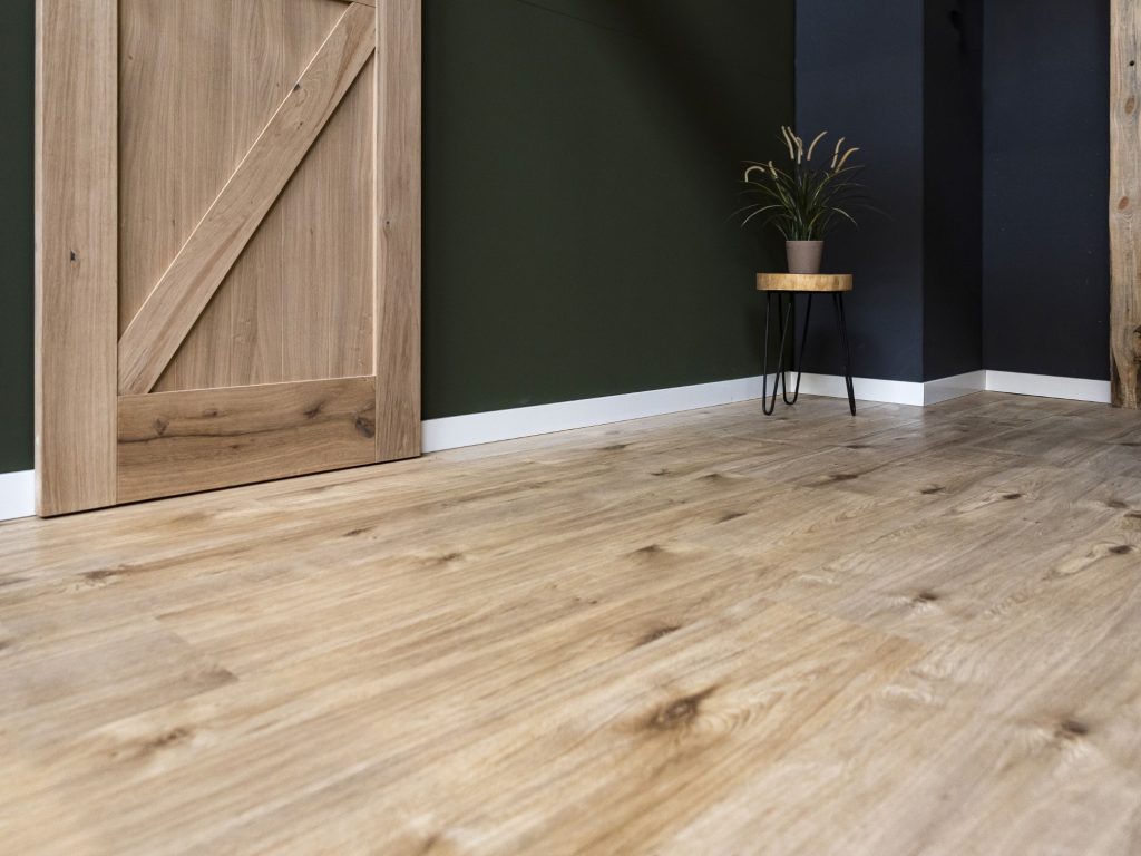 Laminaatvloer in eiken-look en een deel van een eiken barndeur is te zien links op het beeld. De muur is afgewerkt met een witte blokplint en er staat een krukje met zwart stalen poten en rond houten blad met een plantje er op in de hoek. Er zijn verschillende gebruiksklassen van laminaat en deze vloer valt onder klasse 32